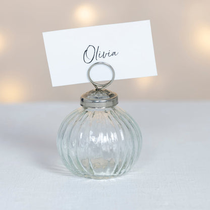 Clear ribbed glass place card holder with silver top and handwritten name card, photographed against a soft neutral background with warm glowing lights.
