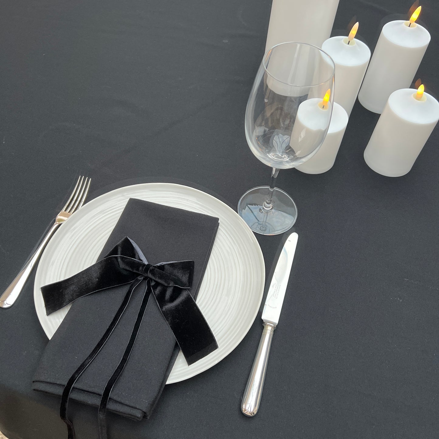Dining table setting with black napkins, silverware, and white candles on a black table linen surface.