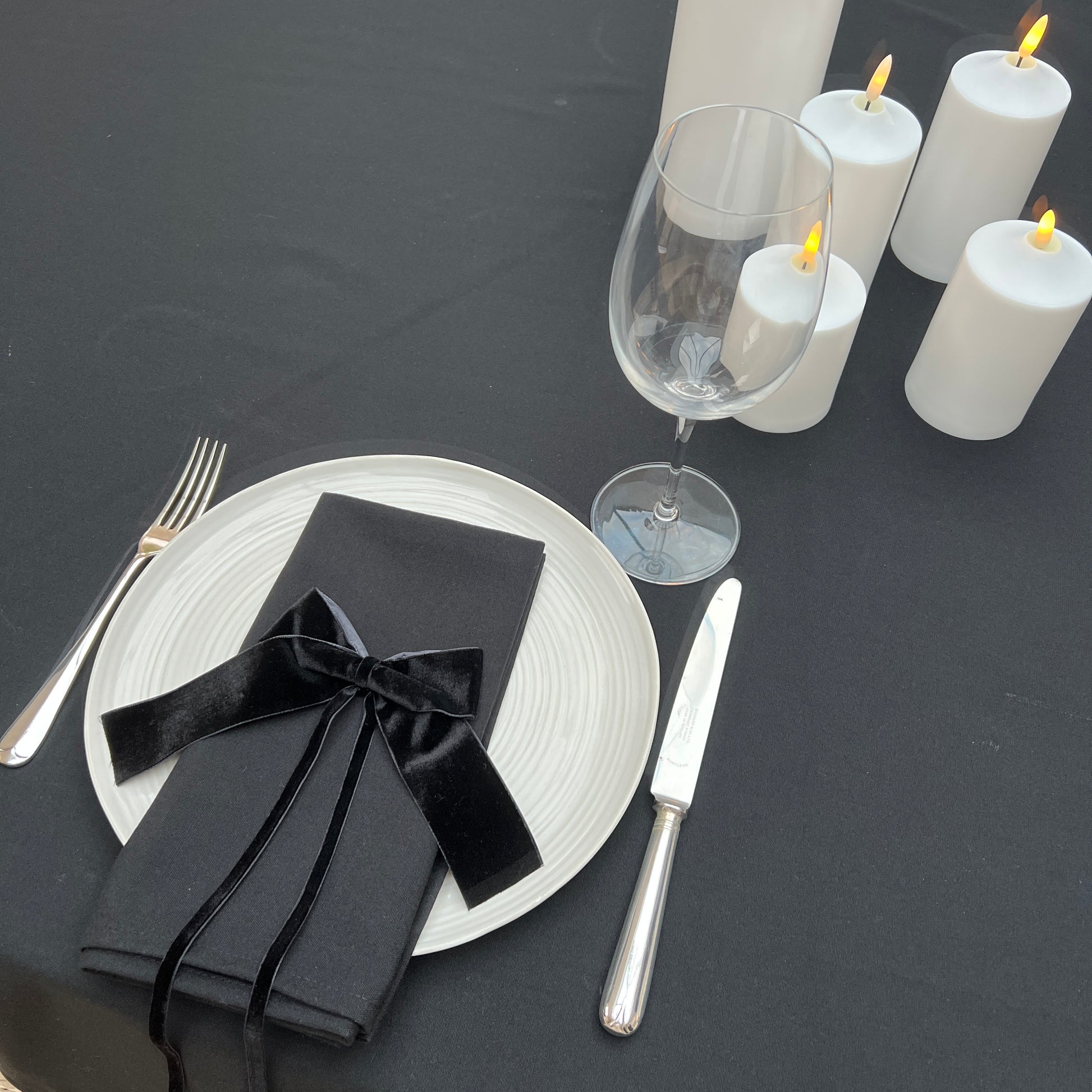 Dining table setting with black napkins, silverware, and white candles on a black table linen surface.