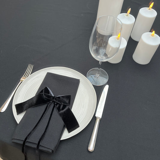 Dining table setting with black napkins, silverware, and white candles on a black table linen surface.