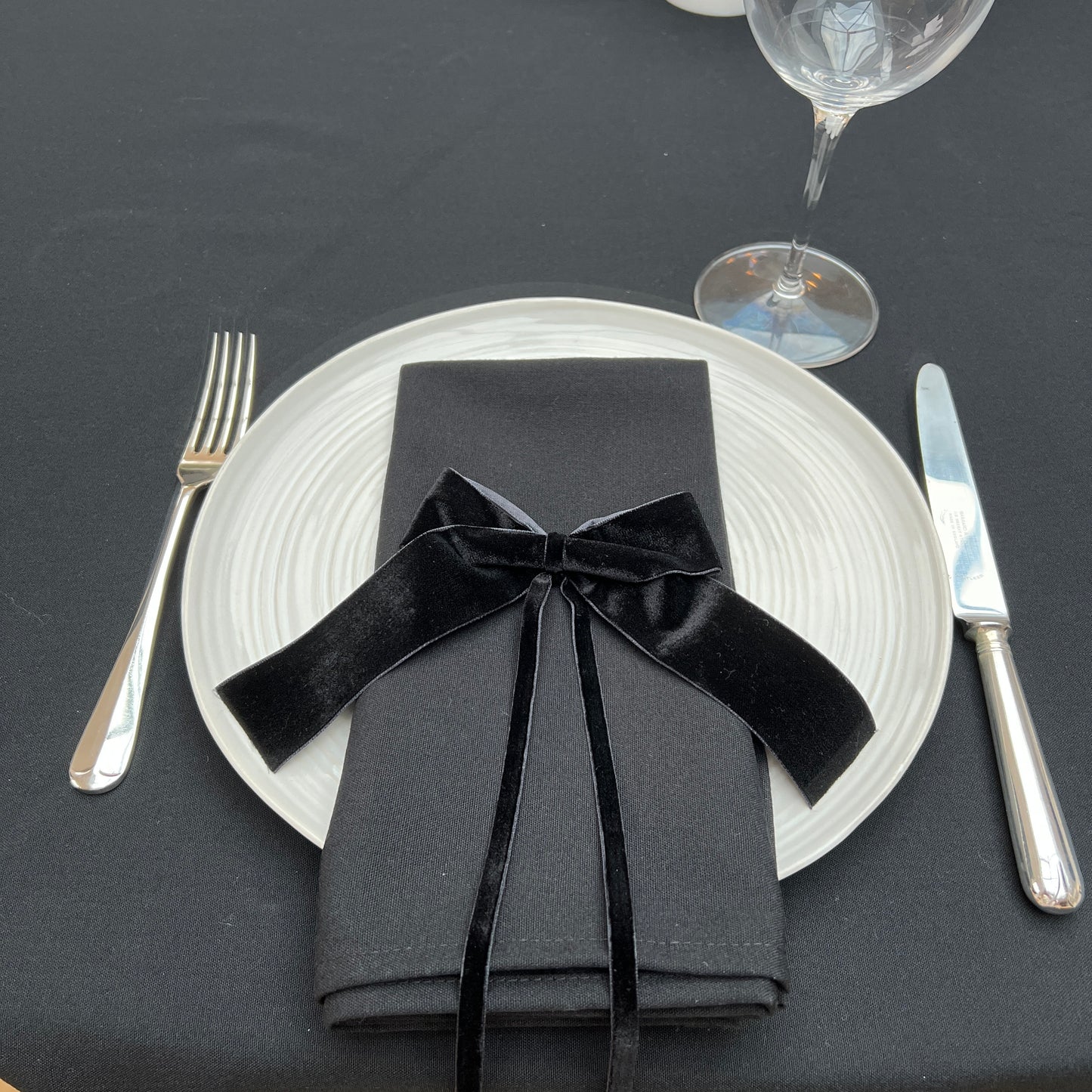 Dinner setting with black napkin tied with a black velvet bow, white plate, silver fork and knife on a black linen tablecloth.