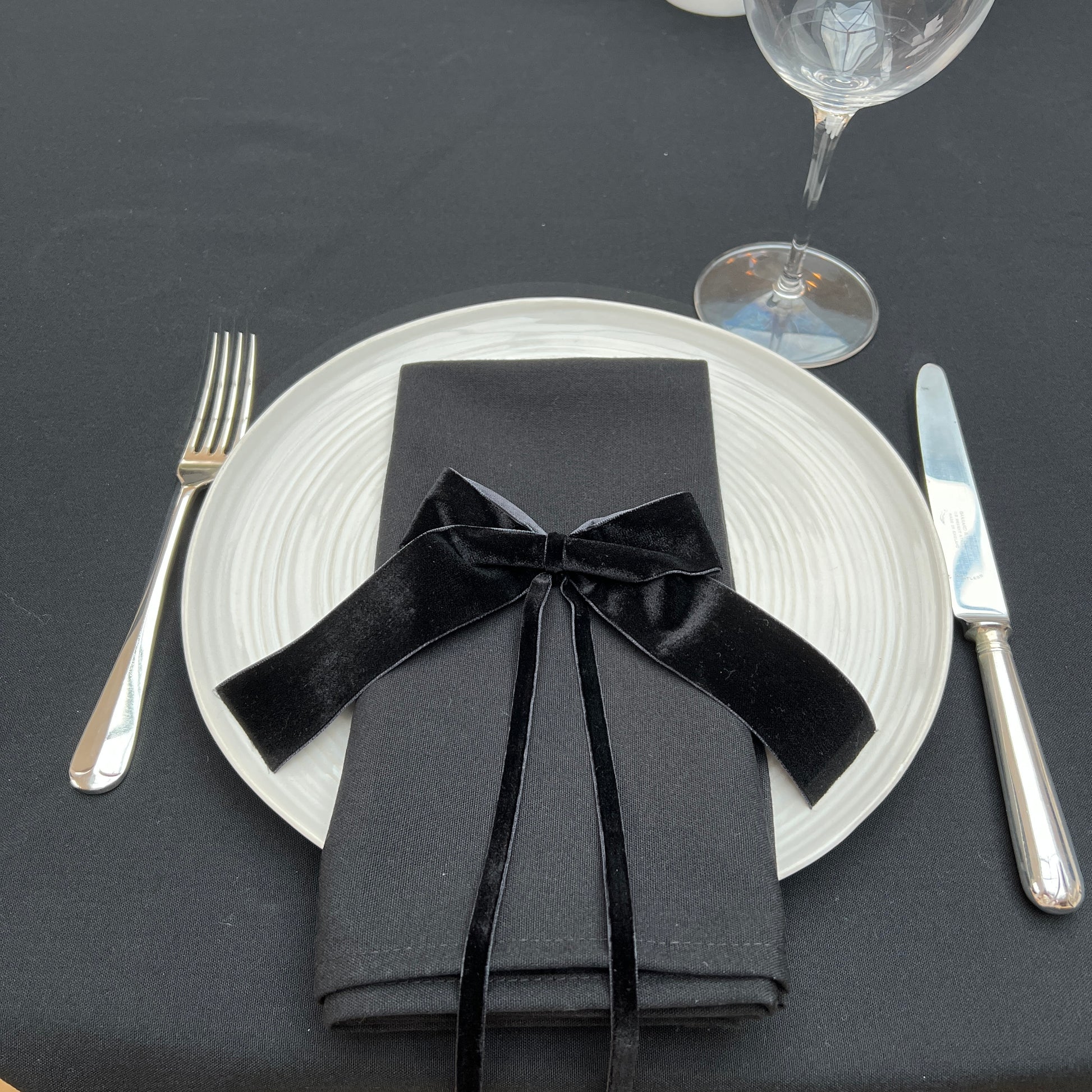 Dinner setting with black napkin tied with a black velvet bow, white plate, silver fork and knife on a black linen tablecloth.
