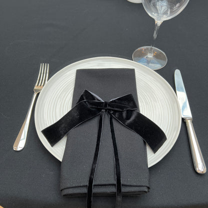 Dinner setting with black napkin tied with a black velvet bow, white plate, silver fork and knife on a black linen tablecloth.