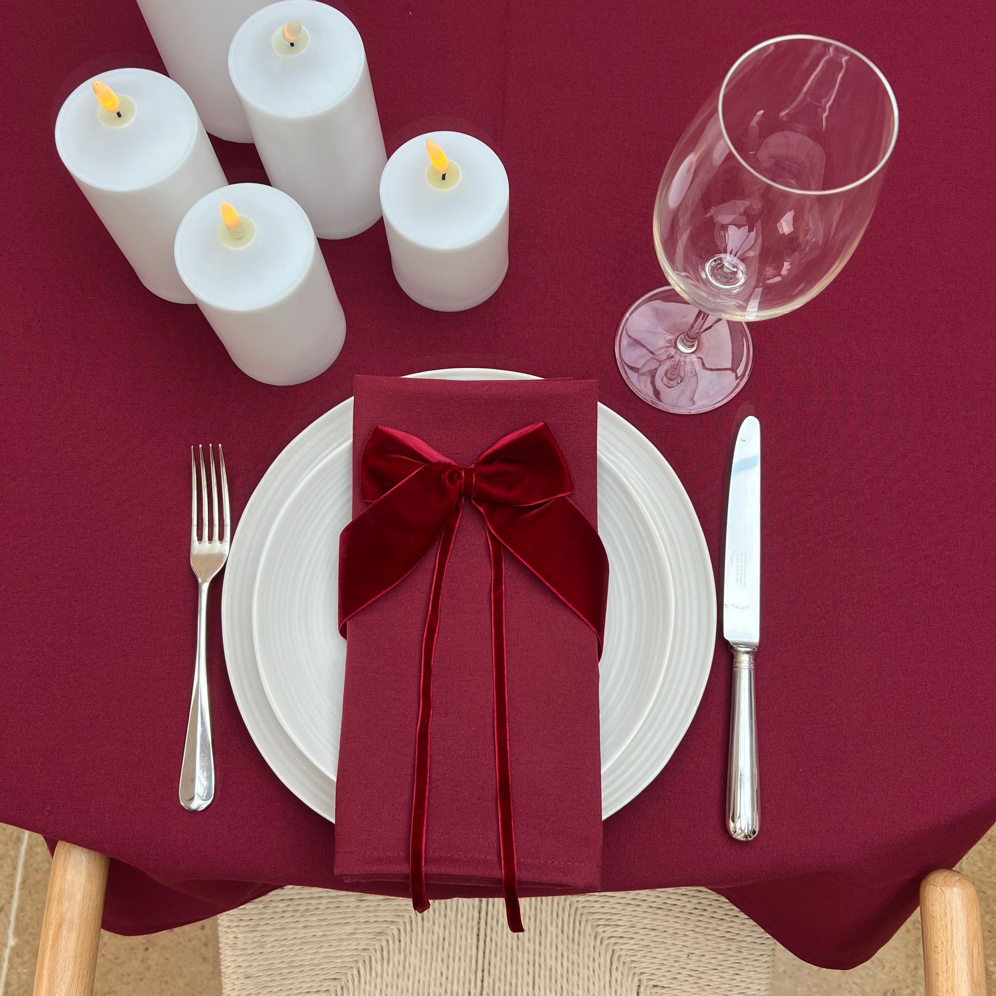 Dining table setting with burgundy napkin, white plates, silverware, and candles on a burgundy tablecloth.