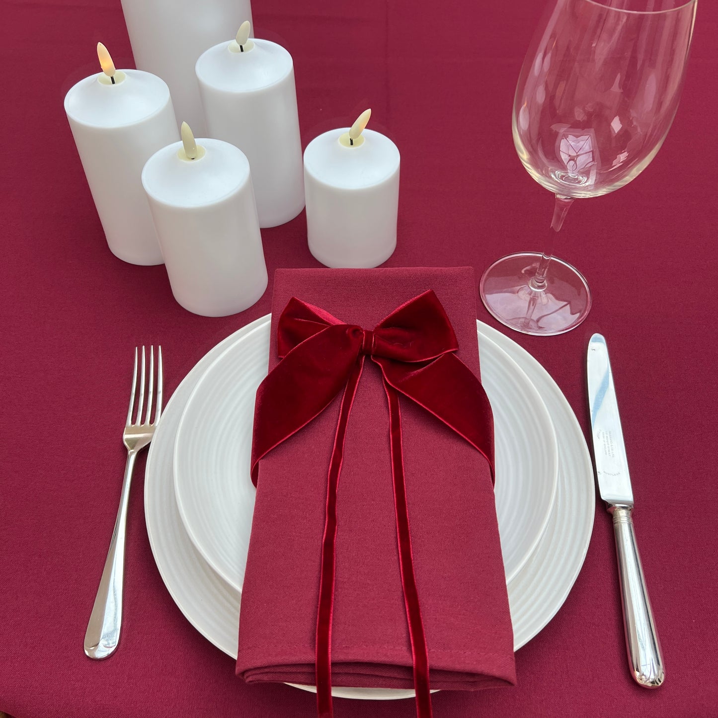 Dinner table setting with burgundy napkin, white candles, and wine glass on a burgundy tablecloth.