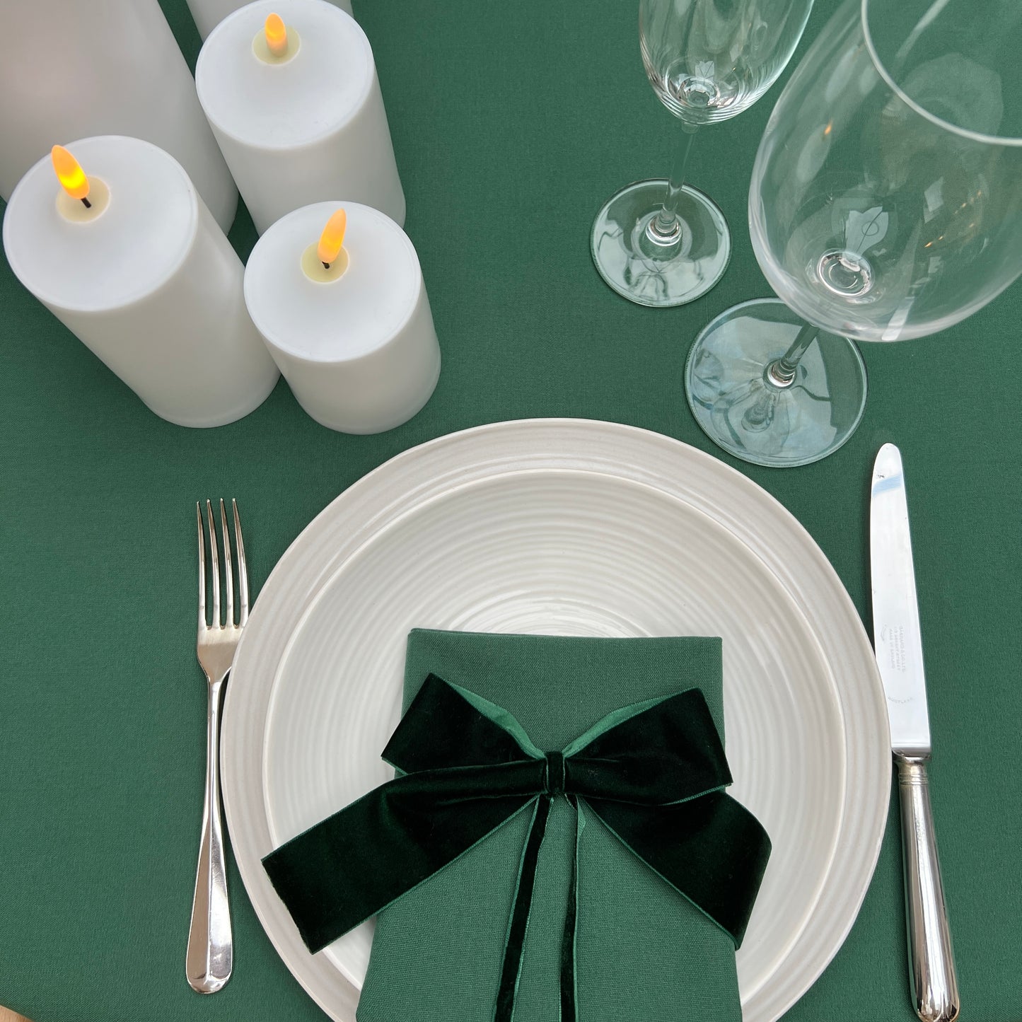 Dining table setting with white plates, silverware, green napkin, and candles on a green tablecloth.