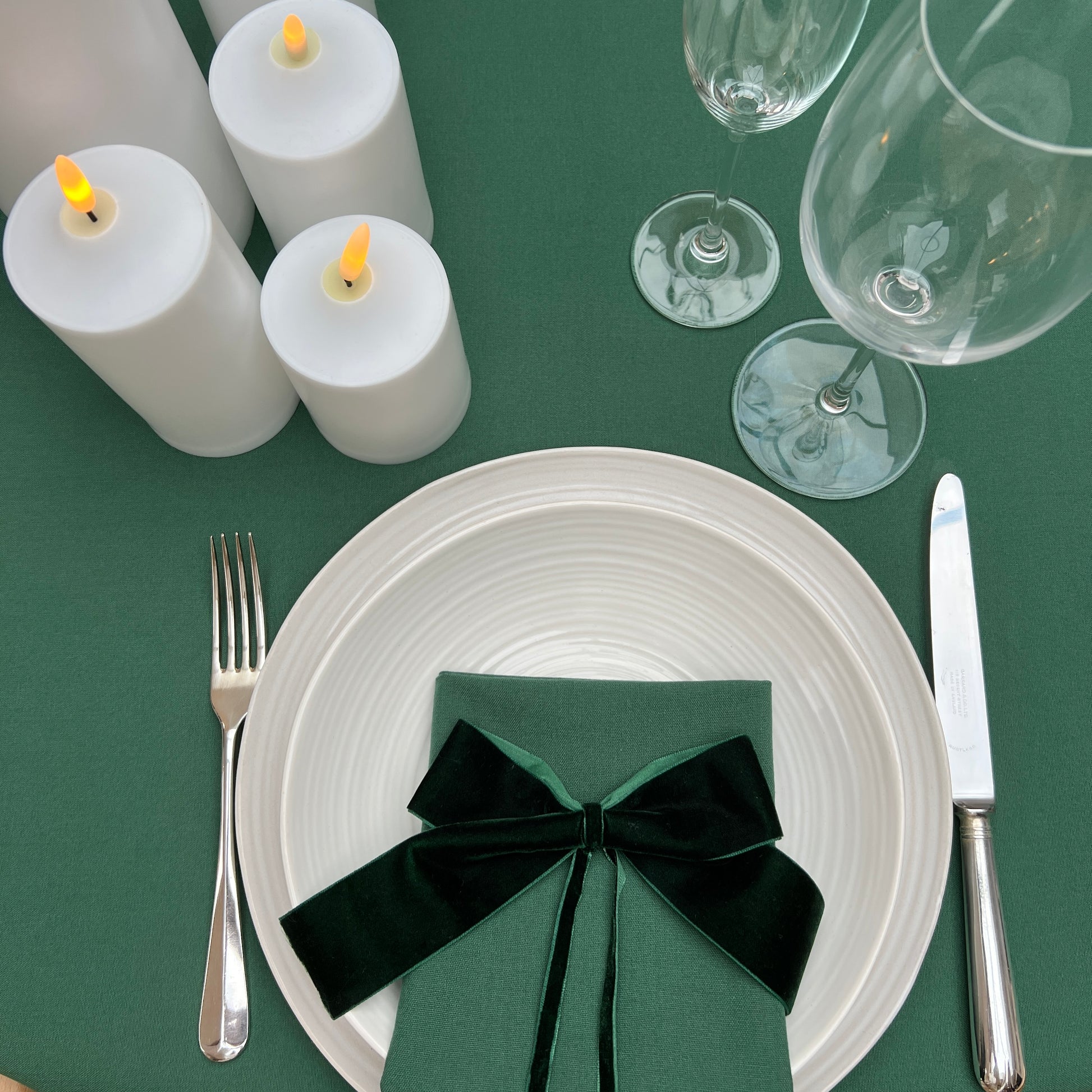 Dining table setting with white plates, silverware, green napkin, and candles on a green tablecloth.