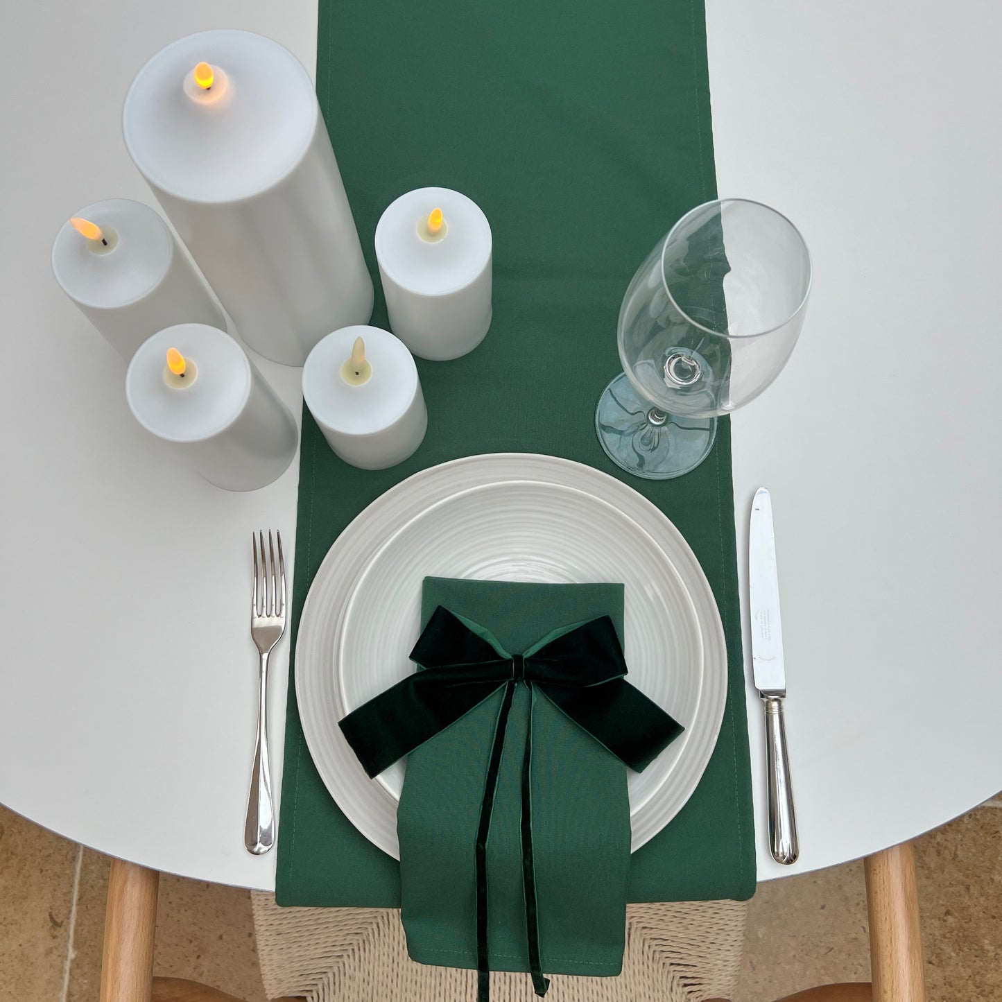 Dining table setting with green runner, white plates, candles, and cutlery on a white surface.