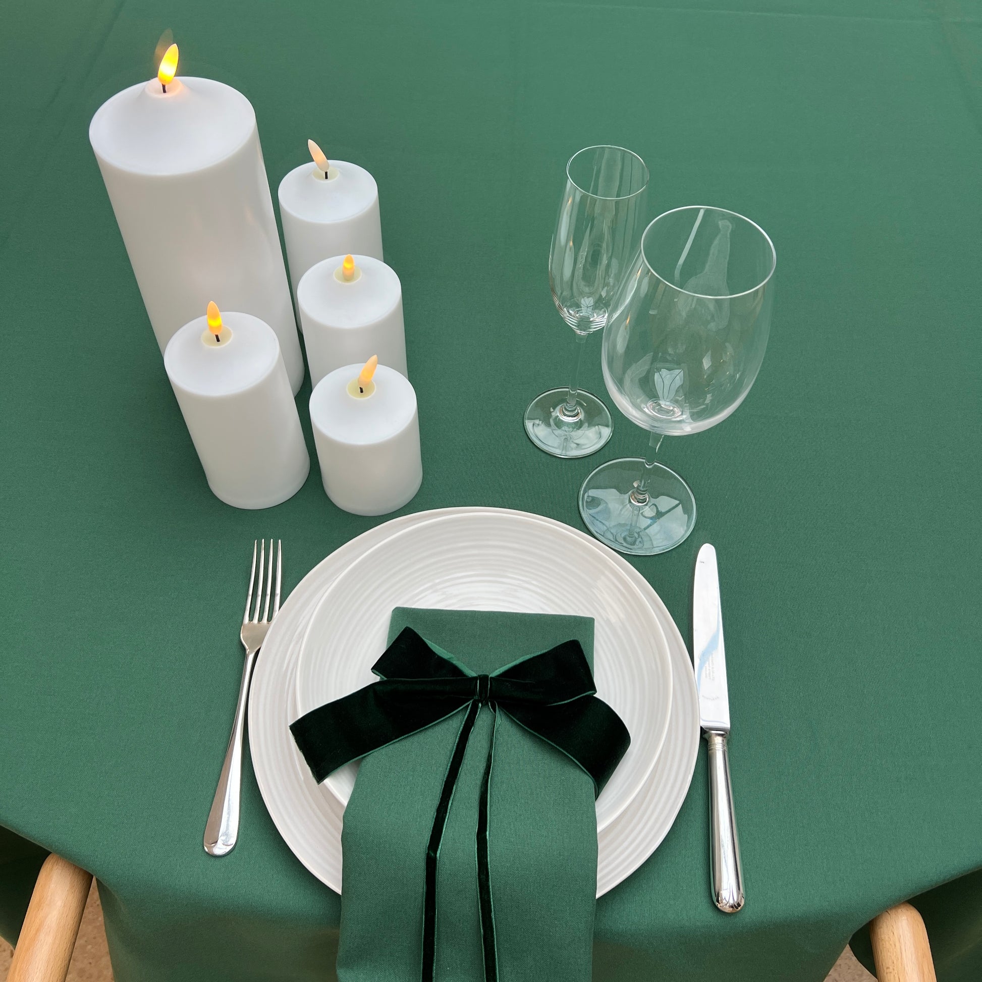 Dining table setting with white candles, glasses, and a green napkin with a dark green velvet bow on a green tablecloth.