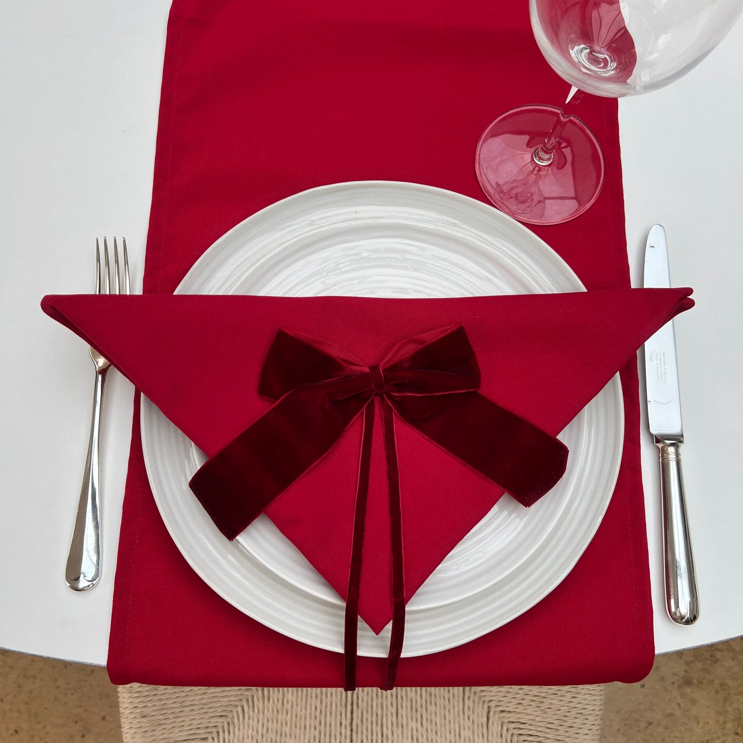 Red napkin with a burgundy velvet bow on a white plate, accompanied by silverware and a glass.