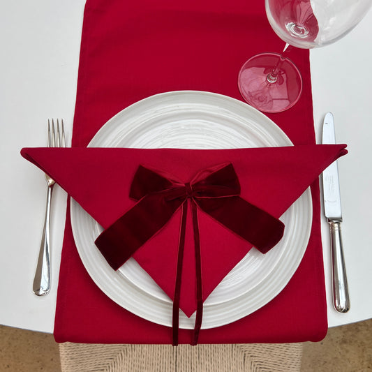 Red napkin with a burgundy velvet bow on a white plate, accompanied by silverware and a glass.