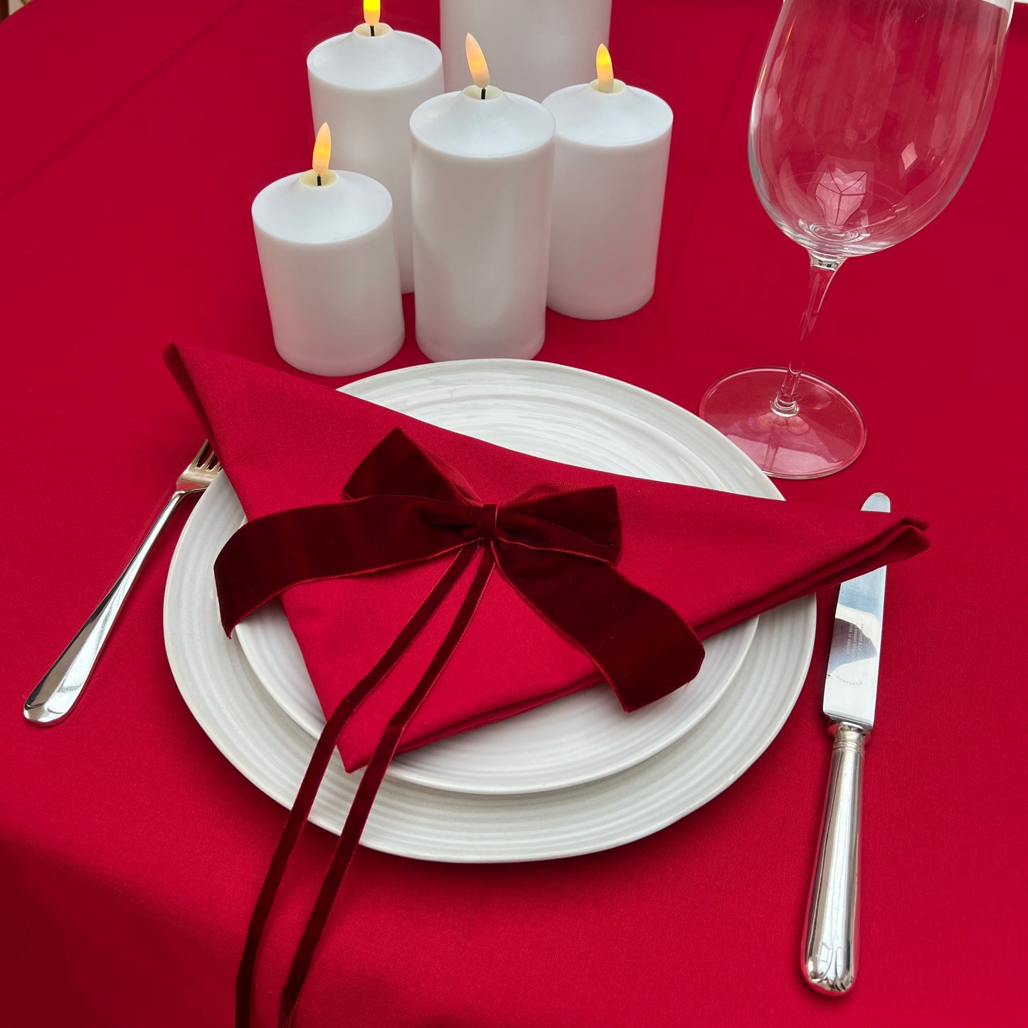 Dinner table setting with red napkin, white plates, candles, and wine glass on a red tablecloth.