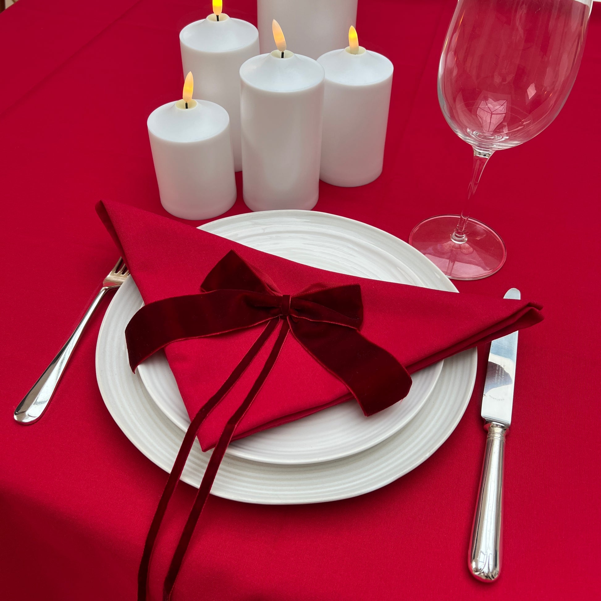 Dinner table setting with red napkin, white plates, candles, and wine glass on a red tablecloth.